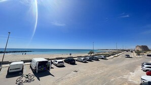 Un parking rempli de voitures et un fourgon blanc, une plage où se trouvent des gens, et un fort historique en arrière-plan.