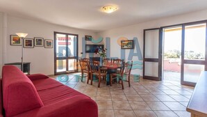 Un'ampia zona giorno con un divano rosso, un tavolo da pranzo con sedie e un televisore fissato alla parete. Si notano quadri appesi e una porta a vetri scorrevoli che dà accesso a una zona esterna.