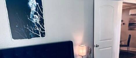 Ein ordentlich gemachtes Bett mit dunklem Kopfteil, weißer Bettwäsche und dekorativen Kissen. Ein Nachttisch mit Lampe und kleiner Statue steht daneben.