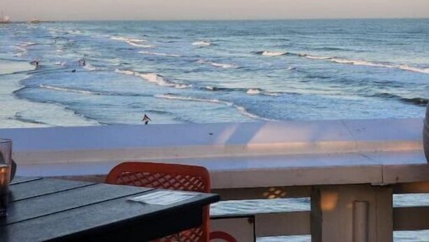 A beachside view with a table and chairs under an umbrella, overlooking the ocean. - Makin’ Waves Cottage- 4 min. to Cruise Ports and half mile from beach (Galveston)