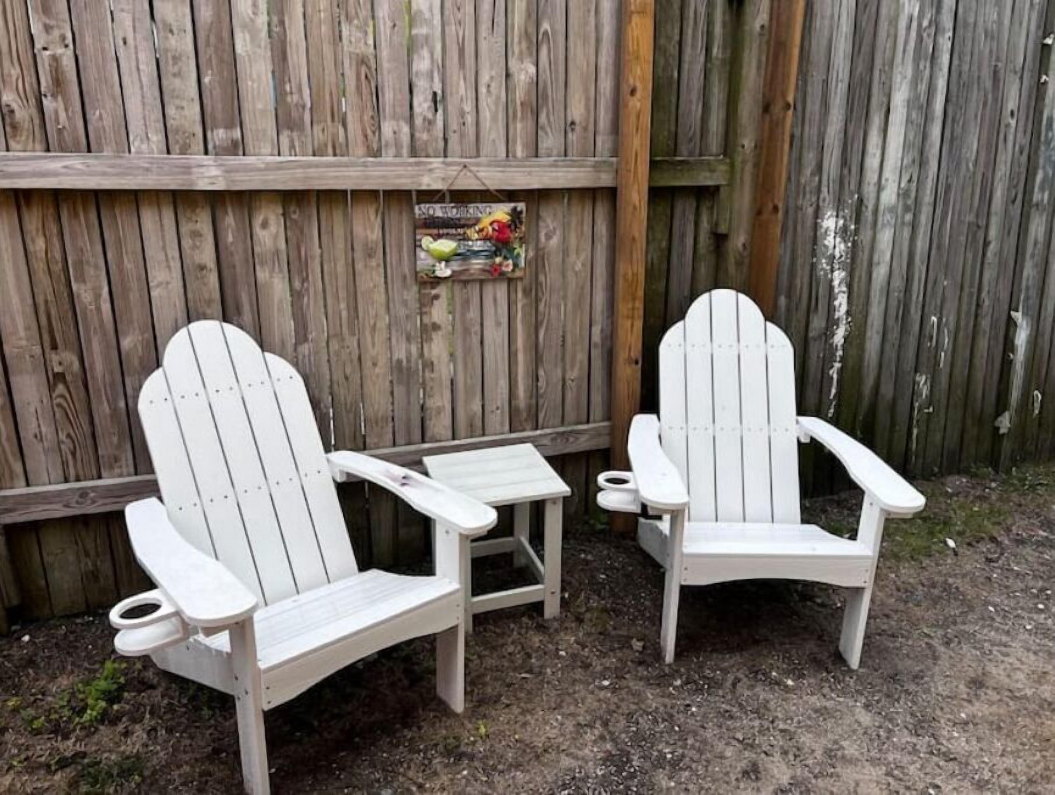 Two Adirondack chairs with a small table in a garden area with a wooden fence in the background.