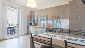 A modern kitchen with wooden cabinets, a dining table with chairs, and a large window. - Apartment Gioia 1 in Marina di Pescoluse (Torre Vado)