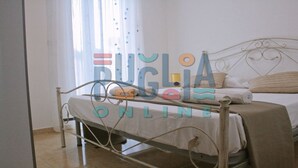 Huoneessa on metallinen sängynrunko, valkoinen päiväpeite ja ikkuna, jossa on läpikuultavat verhot.