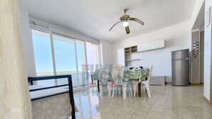 Een ruime kamer met balkon, plafondventilator, een eethoek met stoelen en een koelkast.