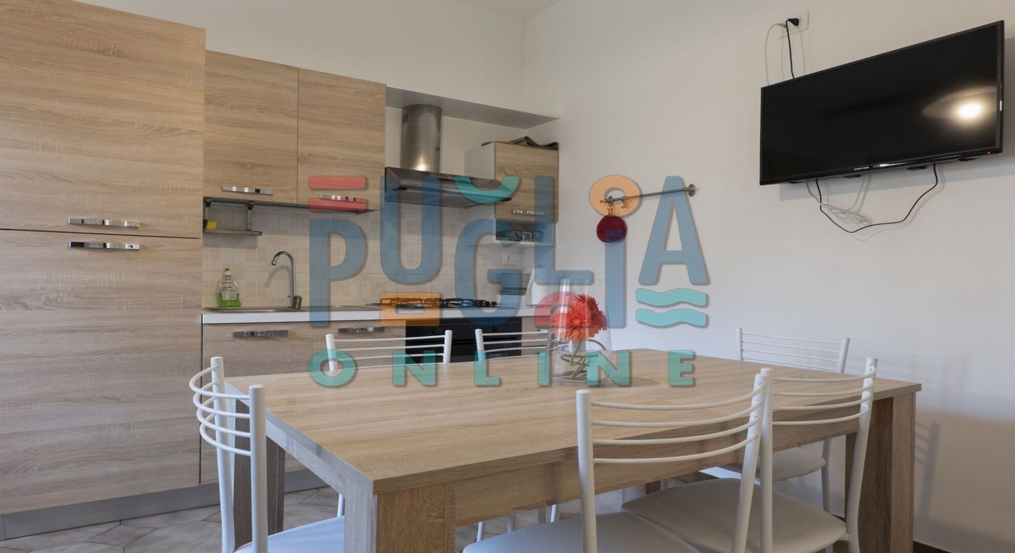 Una cucina moderna con mobili in legno, piano cottura integrato e una TV a schermo piatto fissata alla parete.