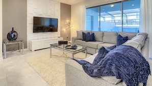 A modern living room with a flat-screen TV, a sofa, a coffee table, and a large window with curtains. - Lovely House 5Beds-4Bath-Private Pool-Club Free (KISSIMMEE)