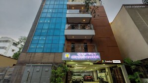 A multi-story building with a prominent blue glass facade, a sign for Hiland Suites, and a parking area with vehicles. - Treebo Hiland Suites (Bengaluru)