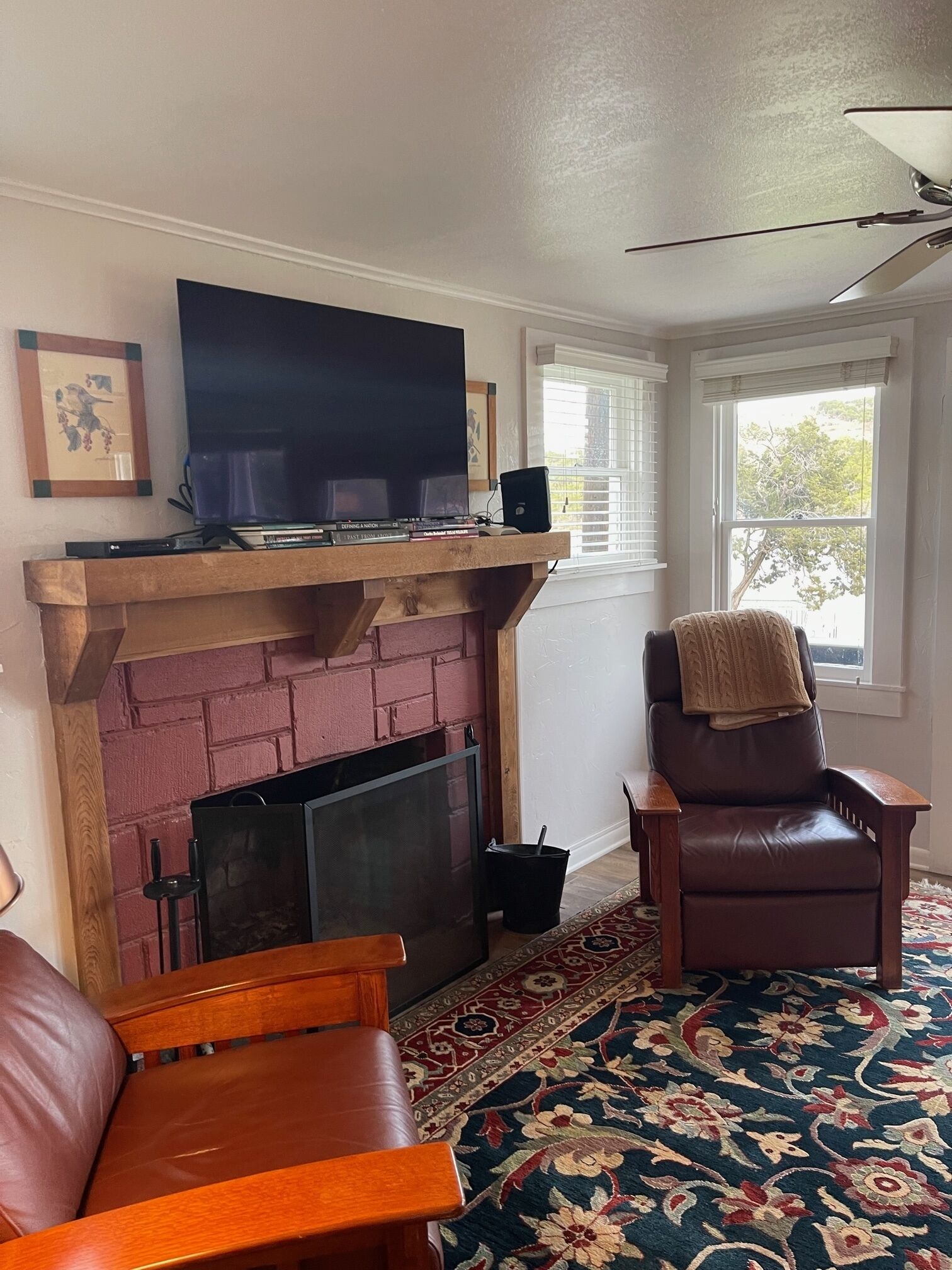 A living room with a brick fireplace, a television on a wooden shelf, a leather armchair, and a patterned rug.