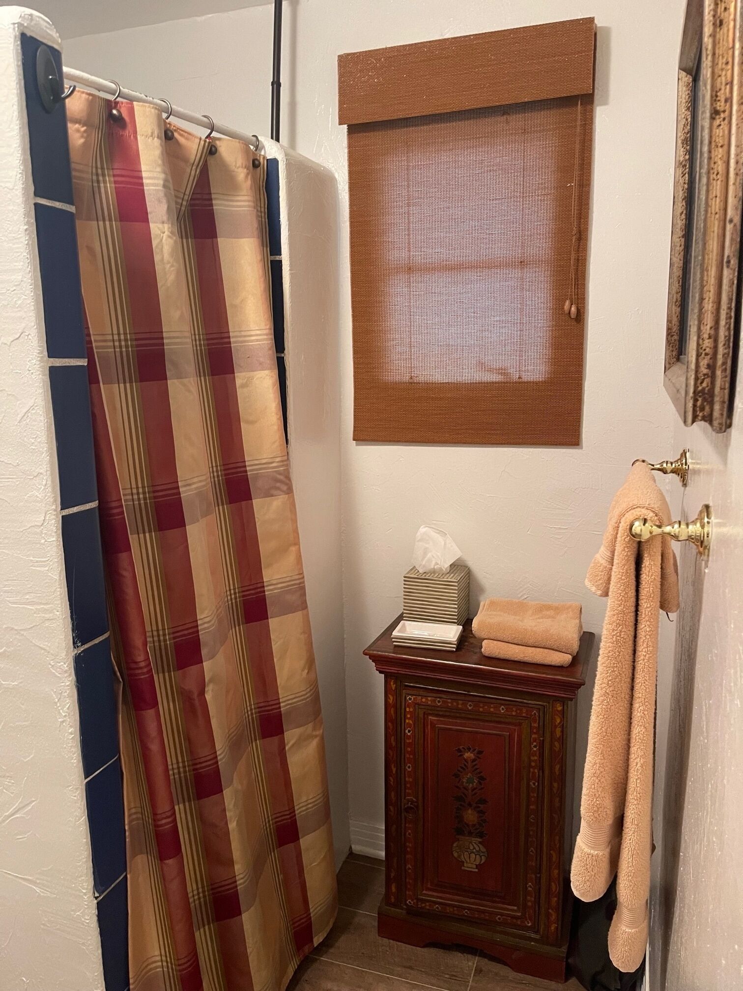 A bathroom with a plaid shower curtain, a small wooden cabinet with a towel, and a framed picture on the wall.