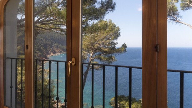 A wooden door with a glass panel and a metal railing, leading to a balcony with a view of the sea and trees.