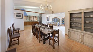 A dining area with a large wooden table set for six, surrounded by chairs. A chandelier hangs from the ceiling, and a cabinet with glassware is visible to the right.