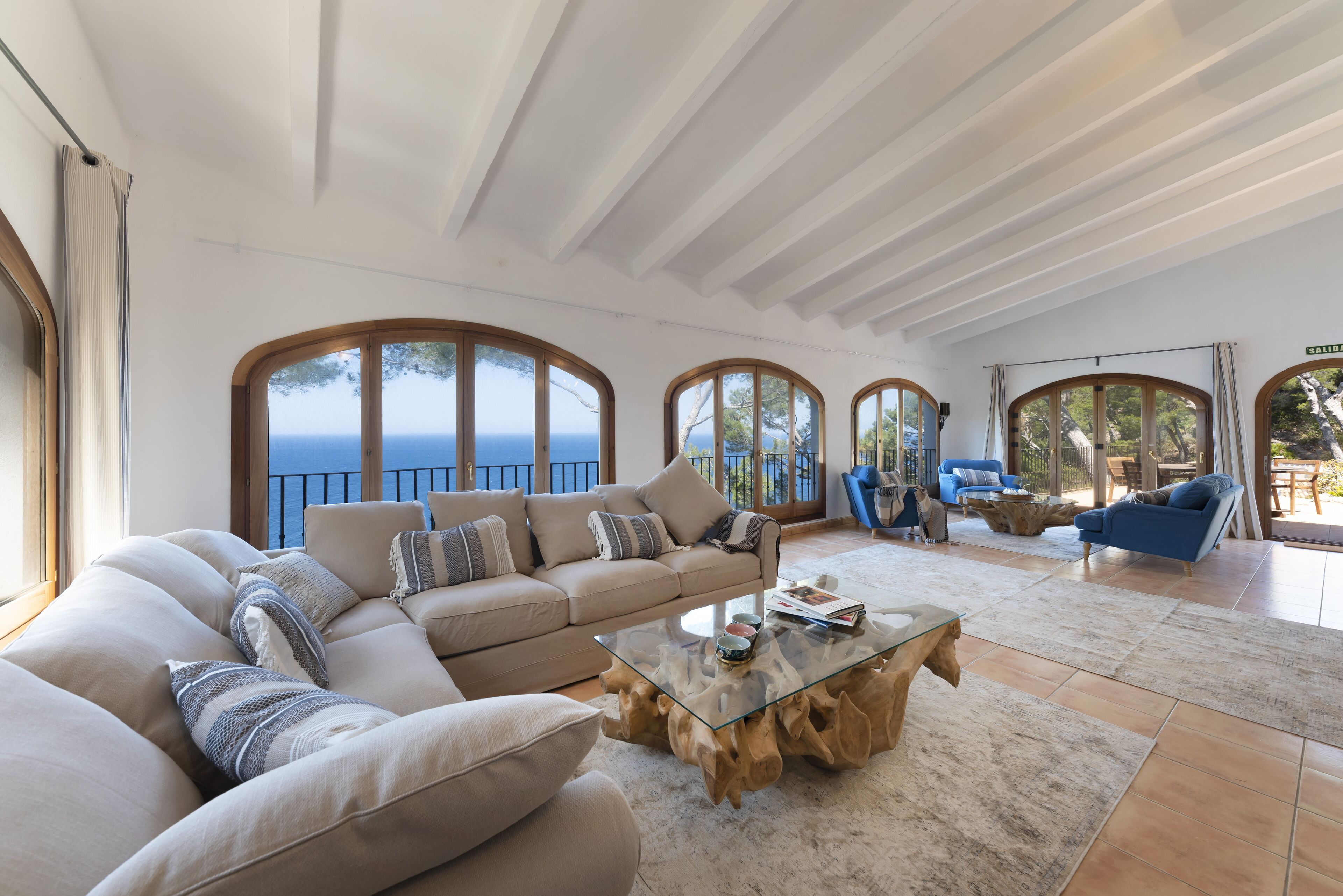 A spacious living room with a large sofa, a glass coffee table, and a view of the sea through arched windows.