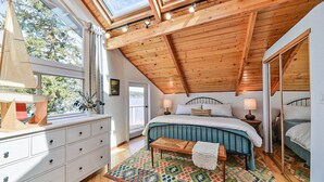 Ein Schlafzimmer mit geneigter Decke, Holzbalken, einem Bett mit blauer Kopfteil, einem Nachttisch mit Lampe, einer Kommode und einem bunten Teppich.