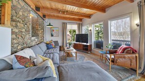 Ein gemütliches Wohnzimmer mit Steinwand, Holzbalkendecke, einem grauen Sofa, einem Ledersessel und einem Flachbildfernseher.