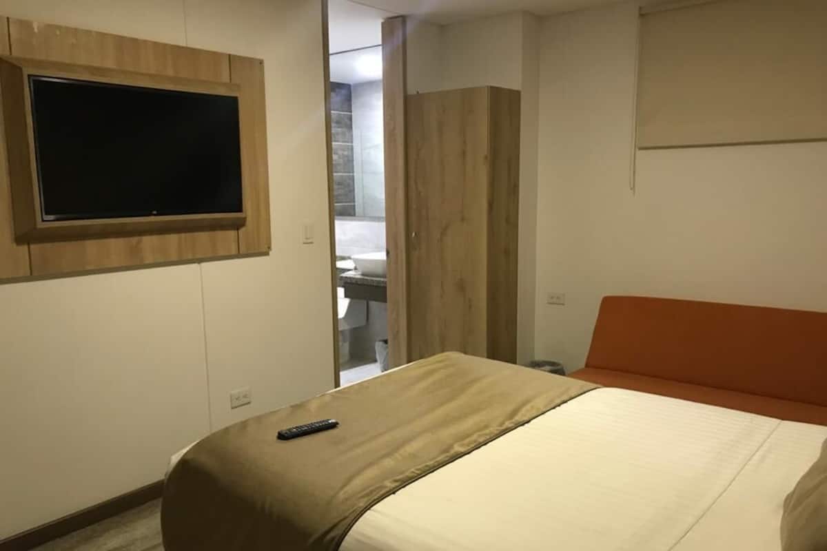 Une chambre d’hôtel avec un lit, un téléviseur fixé au mur et une salle de bain visible à travers une porte ouverte.