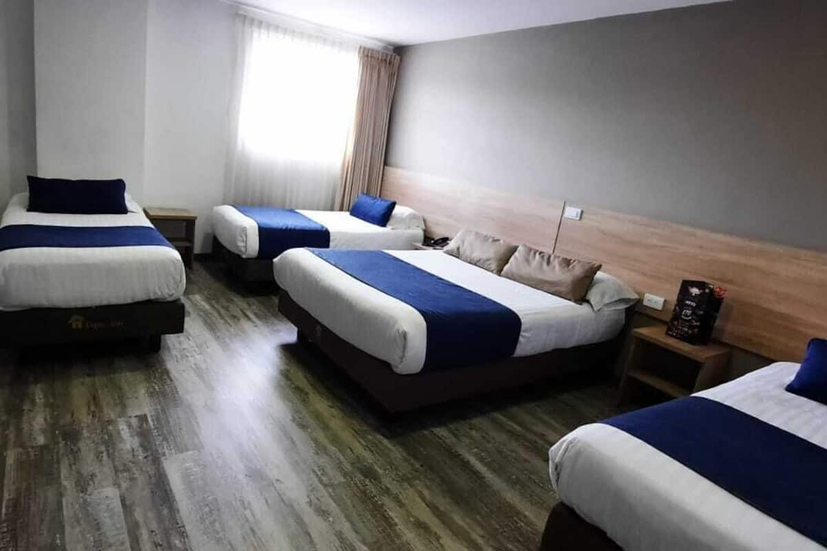 Une chambre d’hôtel avec trois lits alignés, un plancher en bois, une fenêtre avec des rideaux et une table de chevet avec une lampe.
