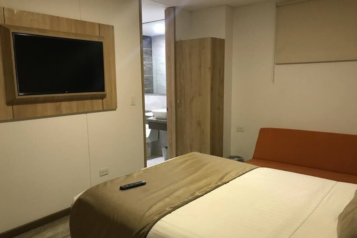 Une chambre d’hôtel avec un lit, un téléviseur fixé au mur, une salle de bain visible à travers une porte ouverte et une armoire en bois.