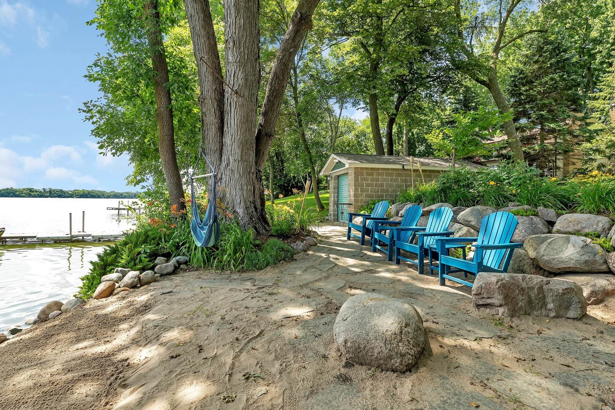 A lakeside cabin with a sandy path leading to blue Adirondack chairs, surrounded by large rocks and trees.