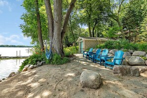 A lakeside cabin with a sandy path leading to blue Adirondack chairs, surrounded by large rocks and trees. - Cozy Lakeside Cabin w/ Fireplace & Great Views (Maple Lake Township)