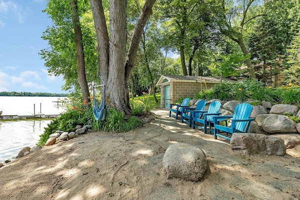 A lakeside cabin with a sandy path leading to blue Adirondack chairs, surrounded by large rocks and trees.