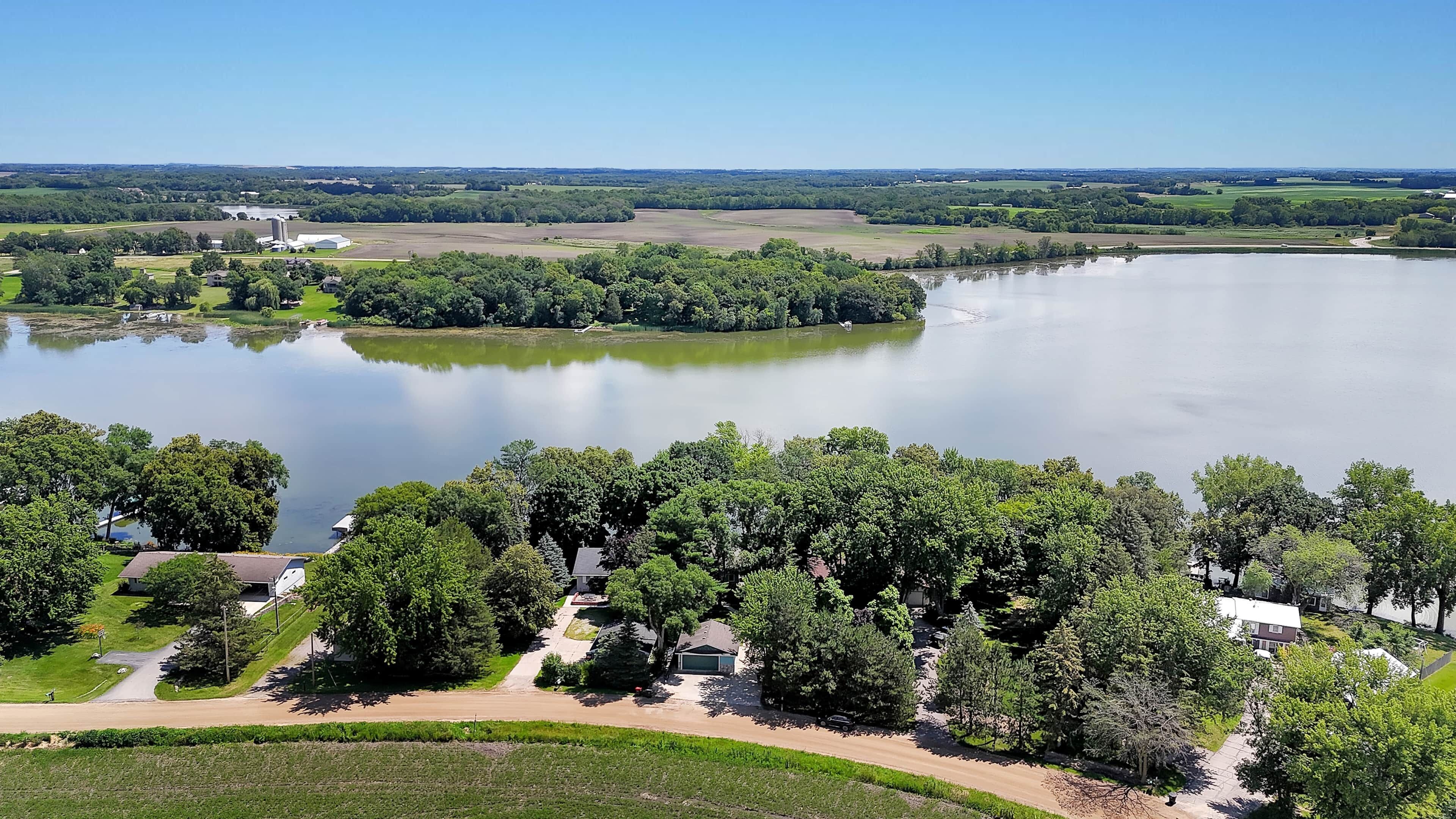 Une propriété située au bord d’un lac, accessible par une route menant à une entrée sécurisée par un portail, et entourée d’une végétation luxuriante et d’une vaste étendue d’eau.