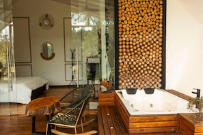 A modern bedroom with a glass door, a wooden wall feature, a large bathtub, and a seating area. - Örök Landscape Hotel (Rionegro)