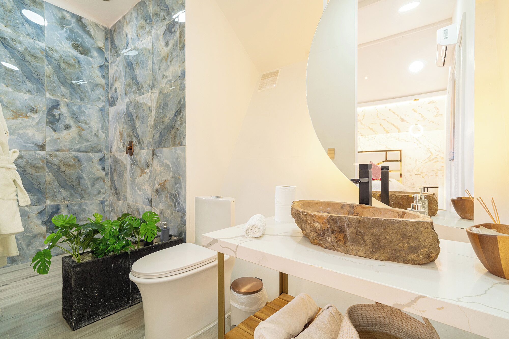 Un baño con pared de mármol, un lavabo con cuenca de piedra grande, inodoro, planta y espejo.
