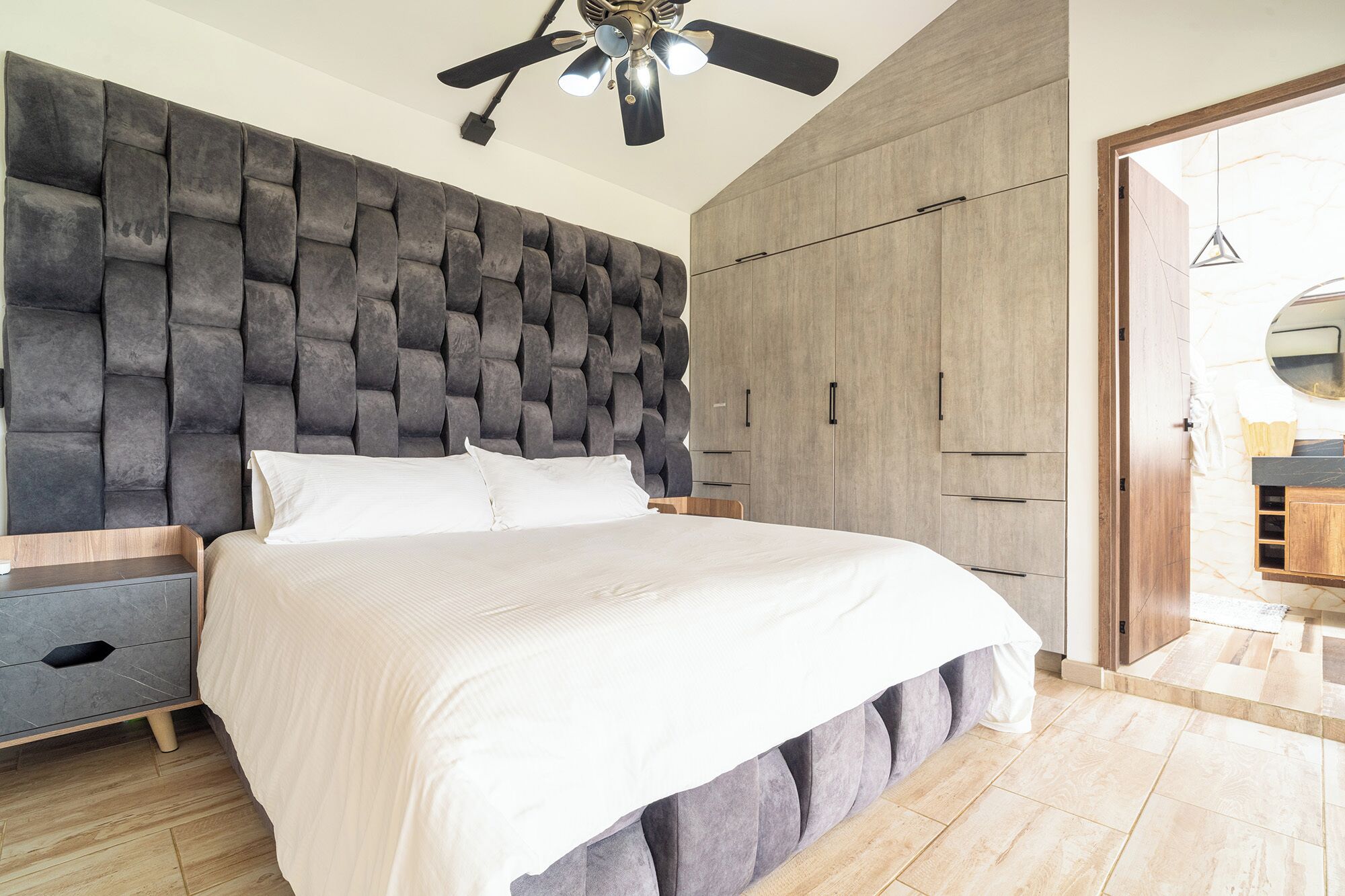 Un dormitorio moderno con cabecera acolchada, cama con ropa de cama blanca, mesita de noche de madera y armario con acabado de madera.