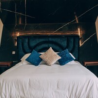 Una cama con ropa de cama blanca, un cabecero oscuro y tres almohadas en azul, beige y azul.