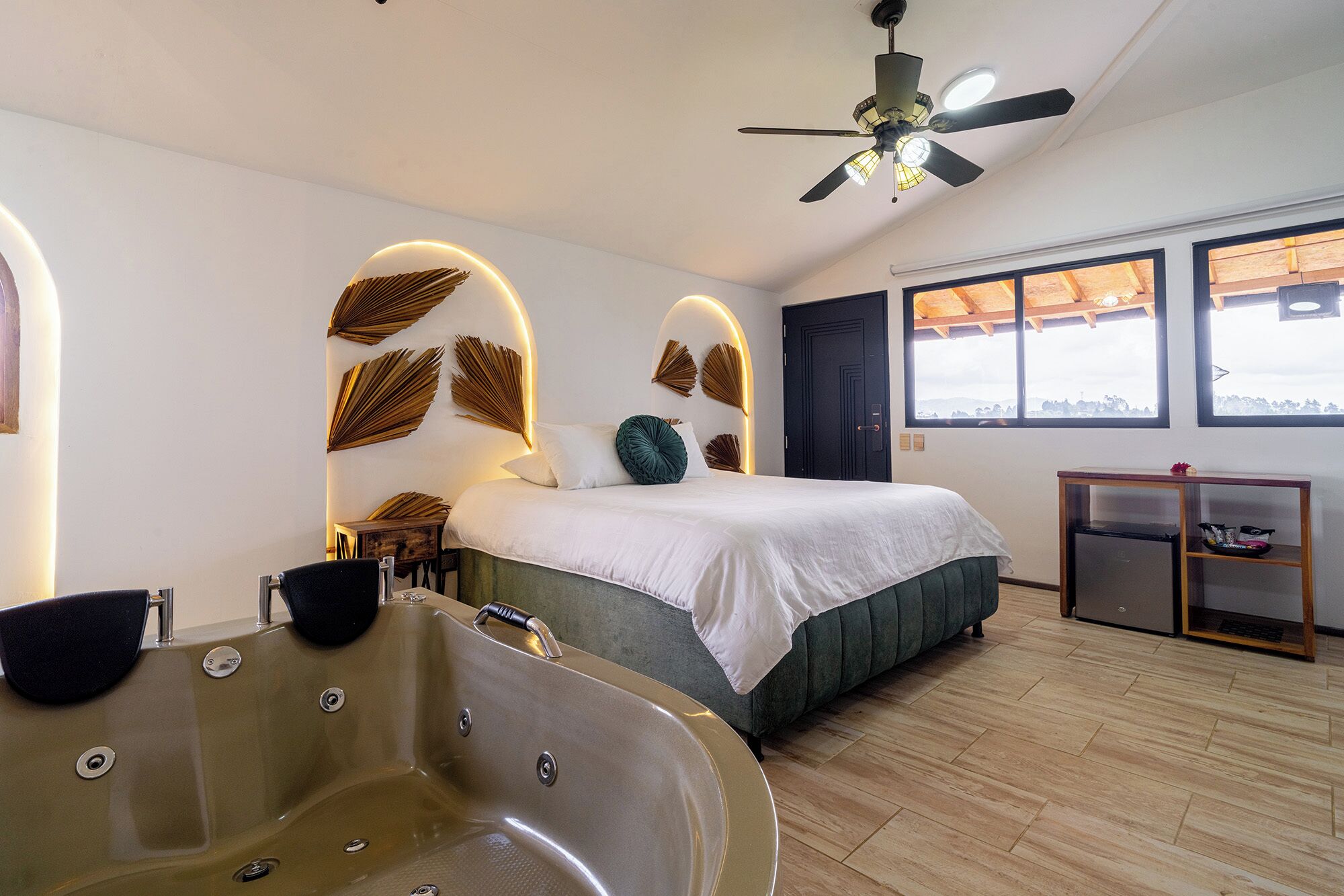 Una habitación de hotel moderna con una cama grande, una zona de estar con una mesa, un ventilador de techo y vistas al exterior.