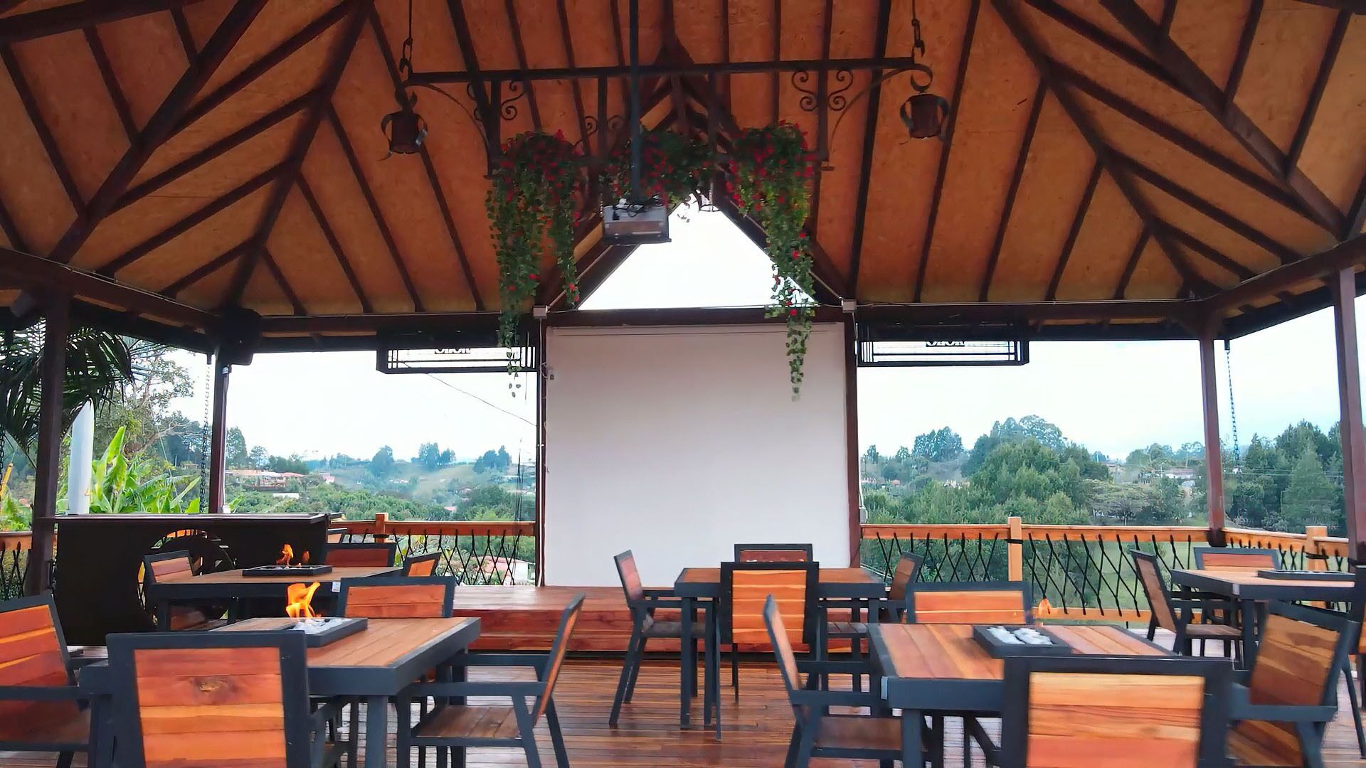 An outdoor dining area with wooden tables and chairs, a white backdrop, and hanging plants.