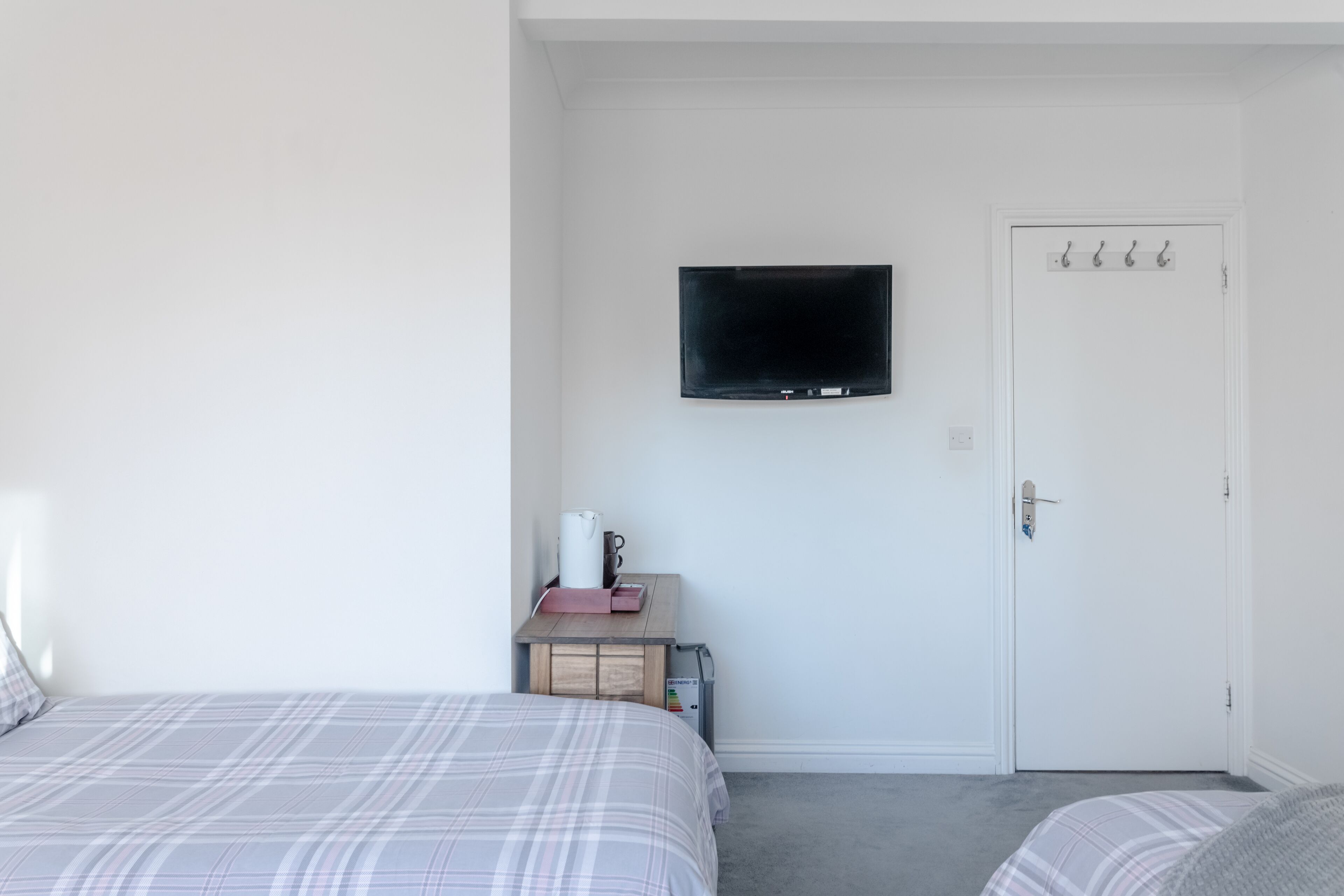 A bedroom with two beds, a television mounted on the wall, a door with hooks, and a small table with a kettle and a pink item.
