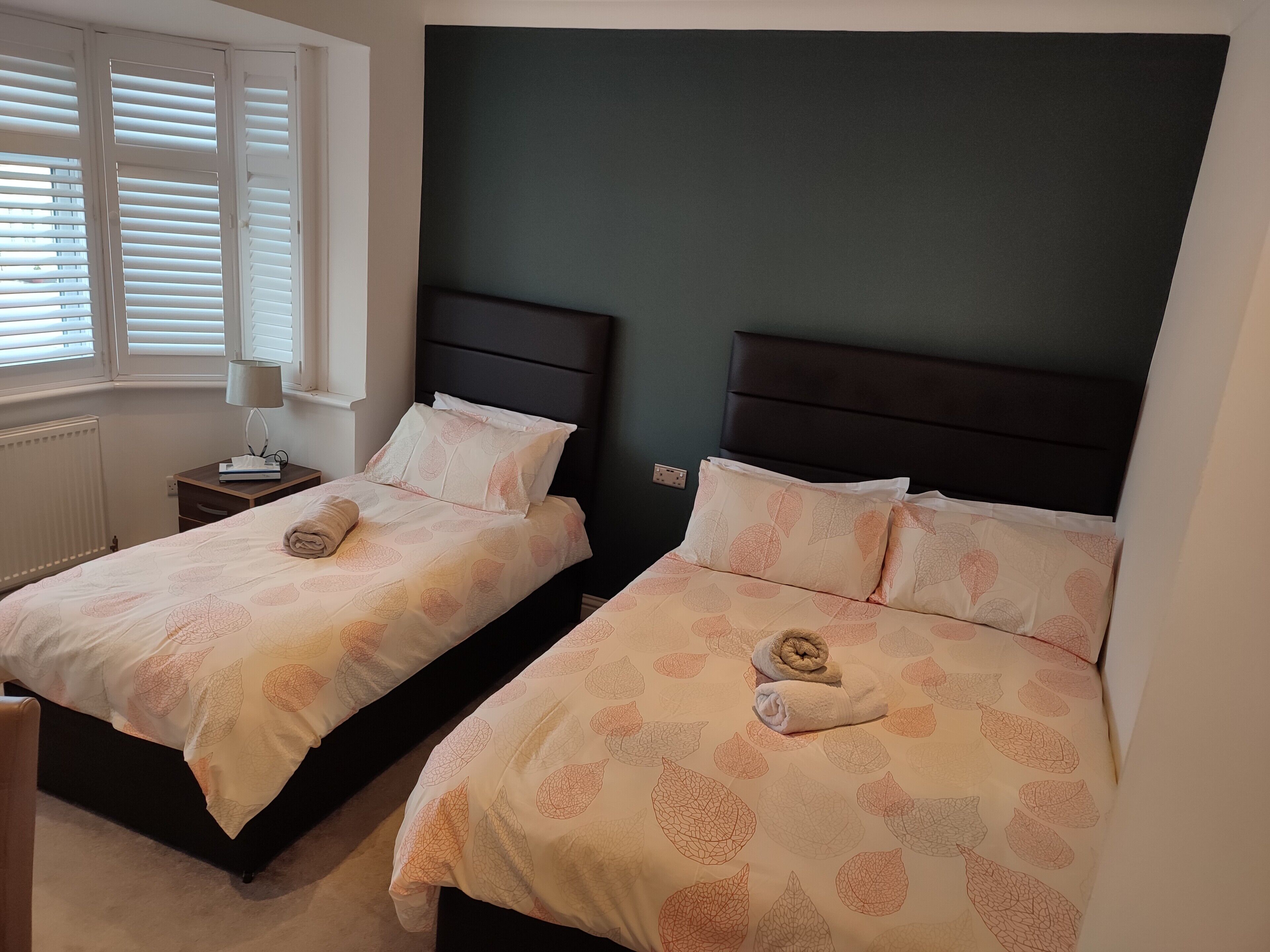 Ein Hotelzimmer mit zwei Betten, jeweils mit dunkelbraunem Kopfteil, weißer Bettwäsche mit Blattmuster, einem Nachttisch mit Lampe und einem Fenster mit Jalousien.