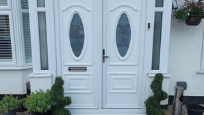 Puerta doble blanca con el número "115" encima, flanqueada por dos ventanas con contraventanas y un pequeño escalón que lleva a la puerta.
