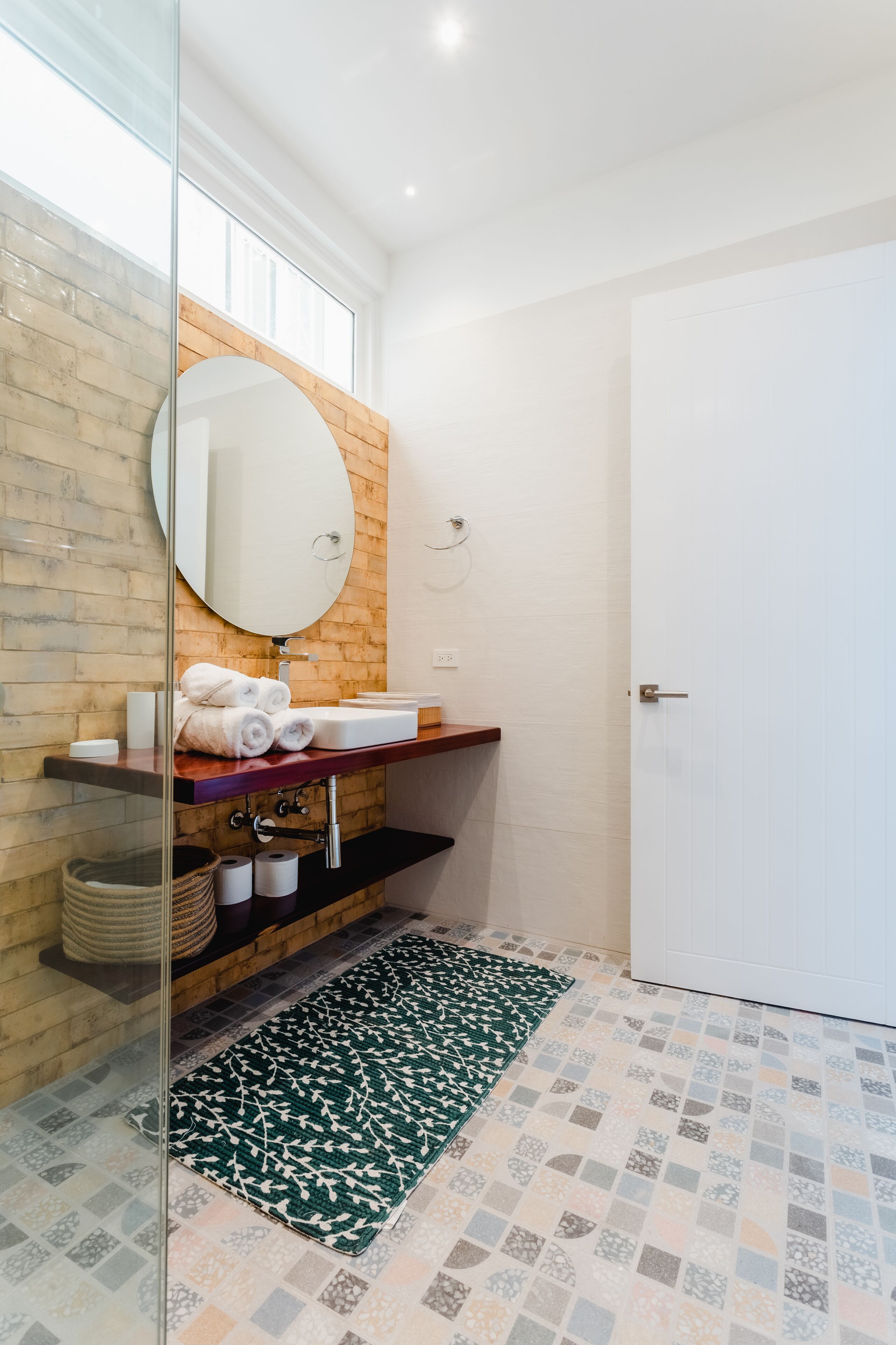 A bathroom with a wooden vanity, a round mirror, a white sink, and a patterned rug on the floor.