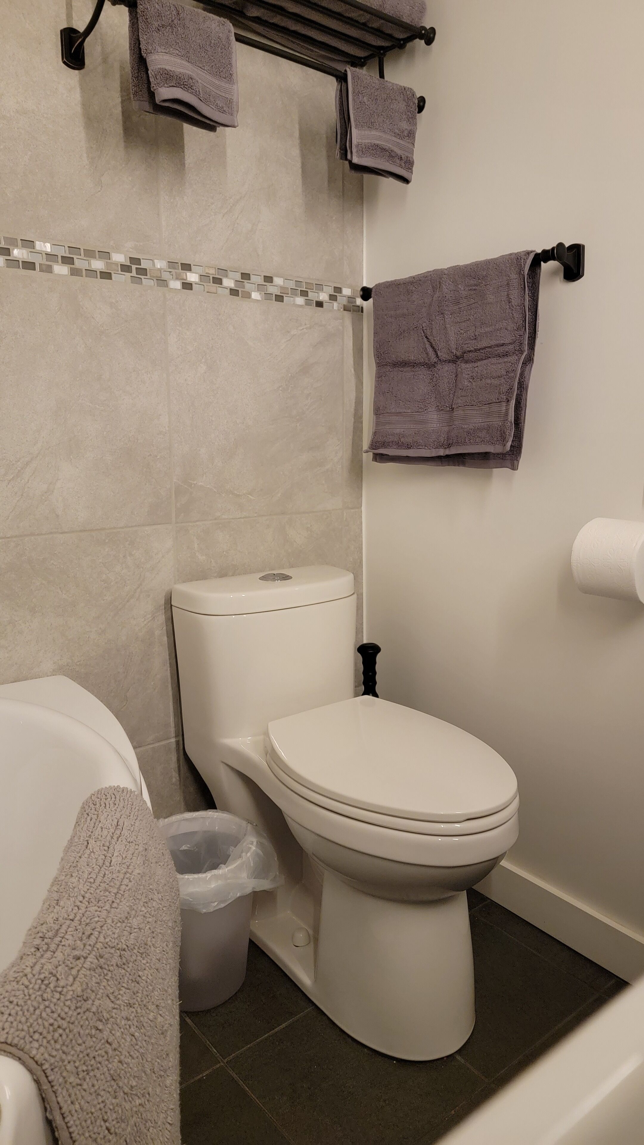 A bathroom with a toilet, a bathtub, a towel rack with towels, and a trash bin.