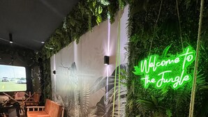 Une pièce avec un plancher en bois, un banc en bois, un canapé et un panneau néon sur lequel on peut lire : « Bienvenue dans la jungle ».