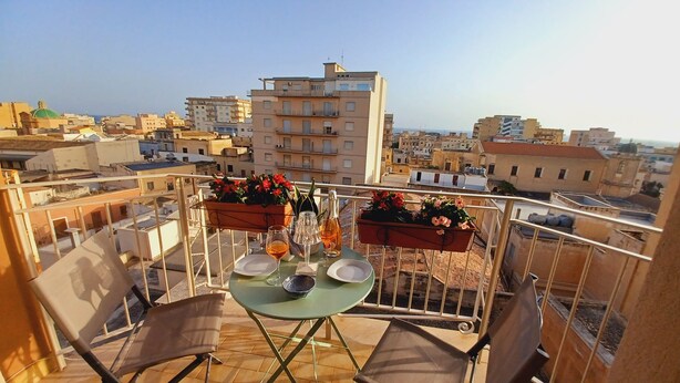 A balcony with a table set for two, potted plants, and a cityscape view. - Family sized trendy flat in the heart of Marsala with balconies and sea view (Marsala)