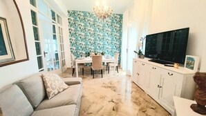 A living room with a sofa, a dining area with a table and chairs, a TV on a cabinet, and a patterned wall. - Family sized trendy flat in the heart of Marsala with balconies and sea view (Marsala)