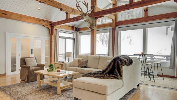 A spacious living room with a high ceiling, wooden beams, a large window, a sofa, a coffee table, and a decorative antler. - Quinn Peaks Retreat House with a hot tub! (Bozeman)