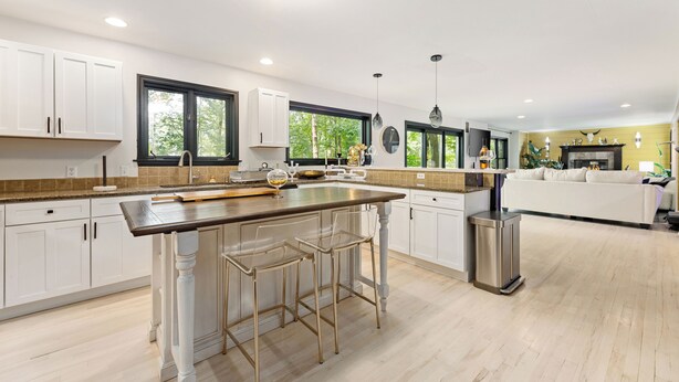 A modern kitchen with white cabinets, a central island with stools, a stainless steel trash can, and a large window providing natural light. - Peaceful Luxury Unique 3 BDR Home w/ Cinema & More (Traverse City)
