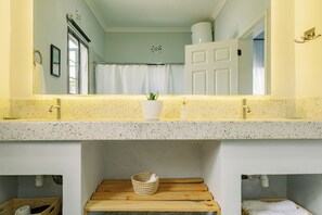 A bathroom with a speckled countertop, a white sink, a mirror, a wooden shelf with a basket, and a door leading to another room. - Sarita Village - Serene Homestay Experience in Arusha's Outskirts. (Arusha)