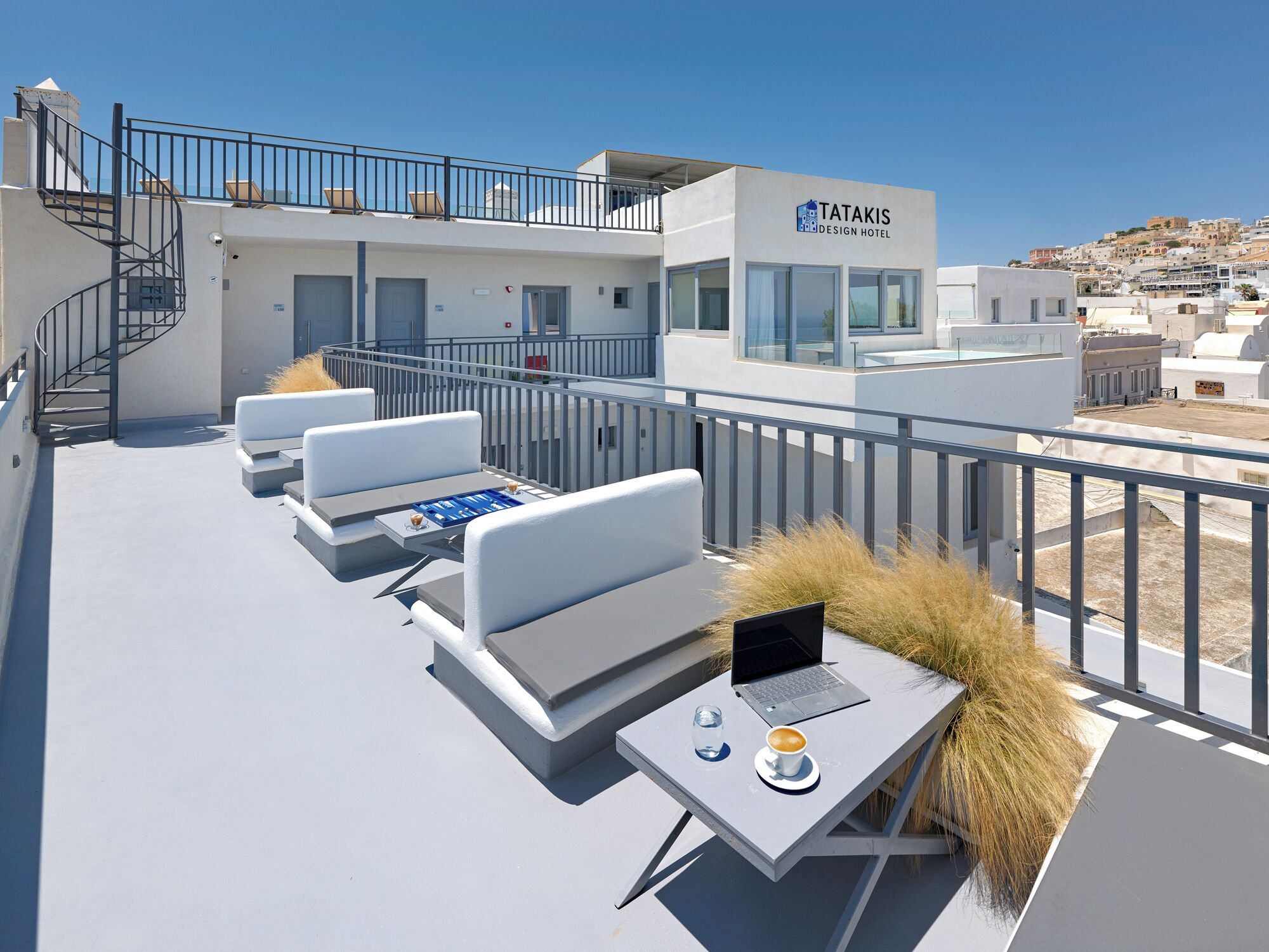 A rooftop terrace with white sofas, a table with a laptop, a coffee cup, and a glass of water.