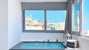 A modern bathroom with a built-in bathtub, a window with curtains, and a view of a cityscape. - Tatakis Design Hotel, Fira (Santorini)
