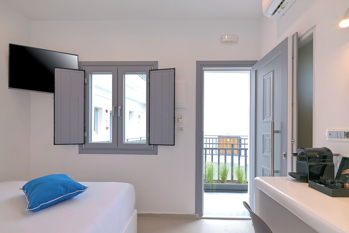 Una habitación de hotel con cama, televisor montado en la pared, cafetera en la encimera y balcón visible a través de la puerta.