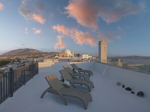 A rooftop terrace with lounge chairs and a view of a coastal town and a church. - Tatakis Design Hotel, Fira (Santorini)