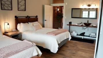 A bedroom with two beds, a wooden headboard, a bedside table with a lamp, a brick wall, a mirror, a shelf with toiletries, and a wooden bench.
