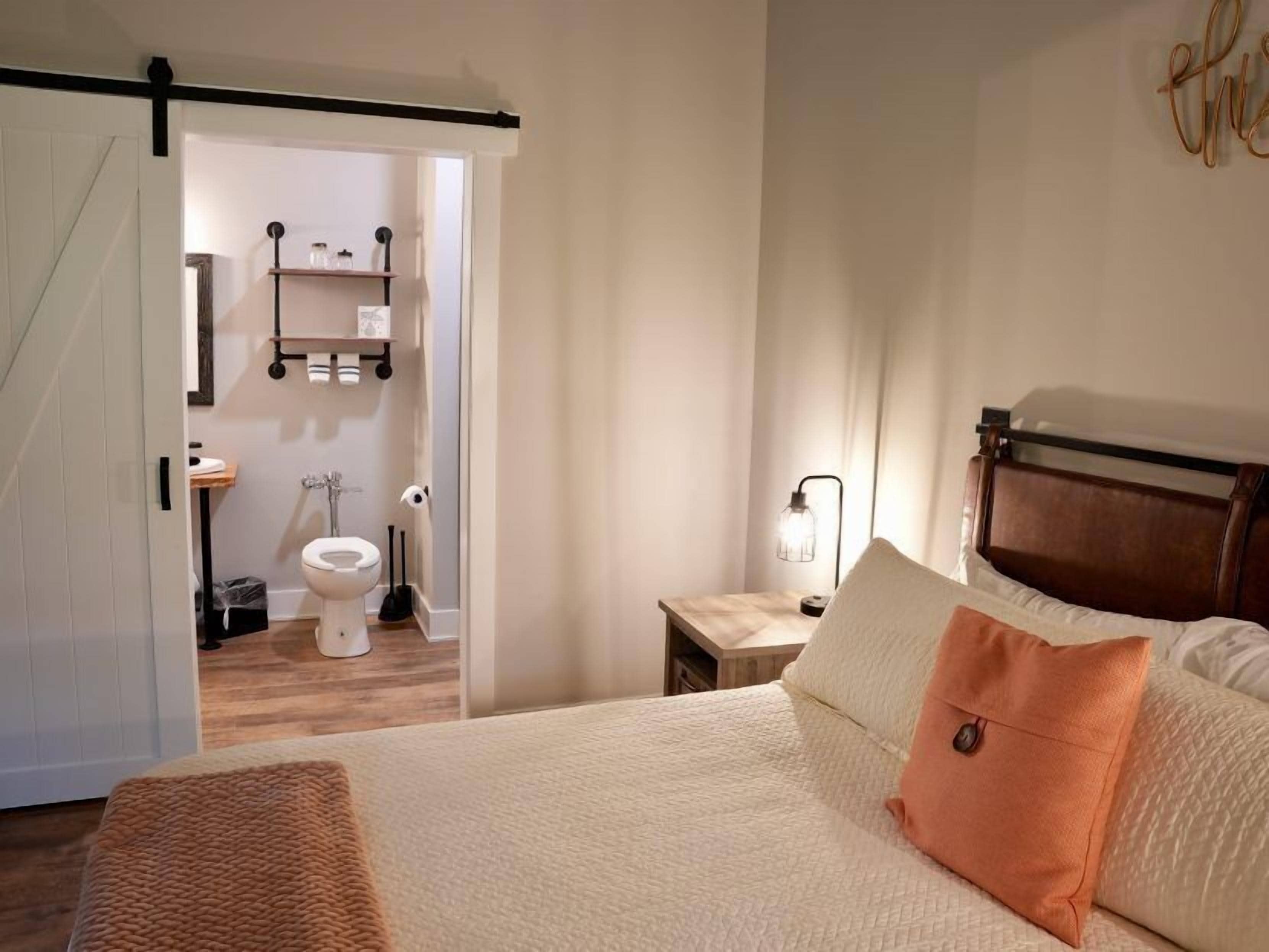 A bedroom with a bed, a nightstand, a bedside lamp, and a view into the bathroom.
