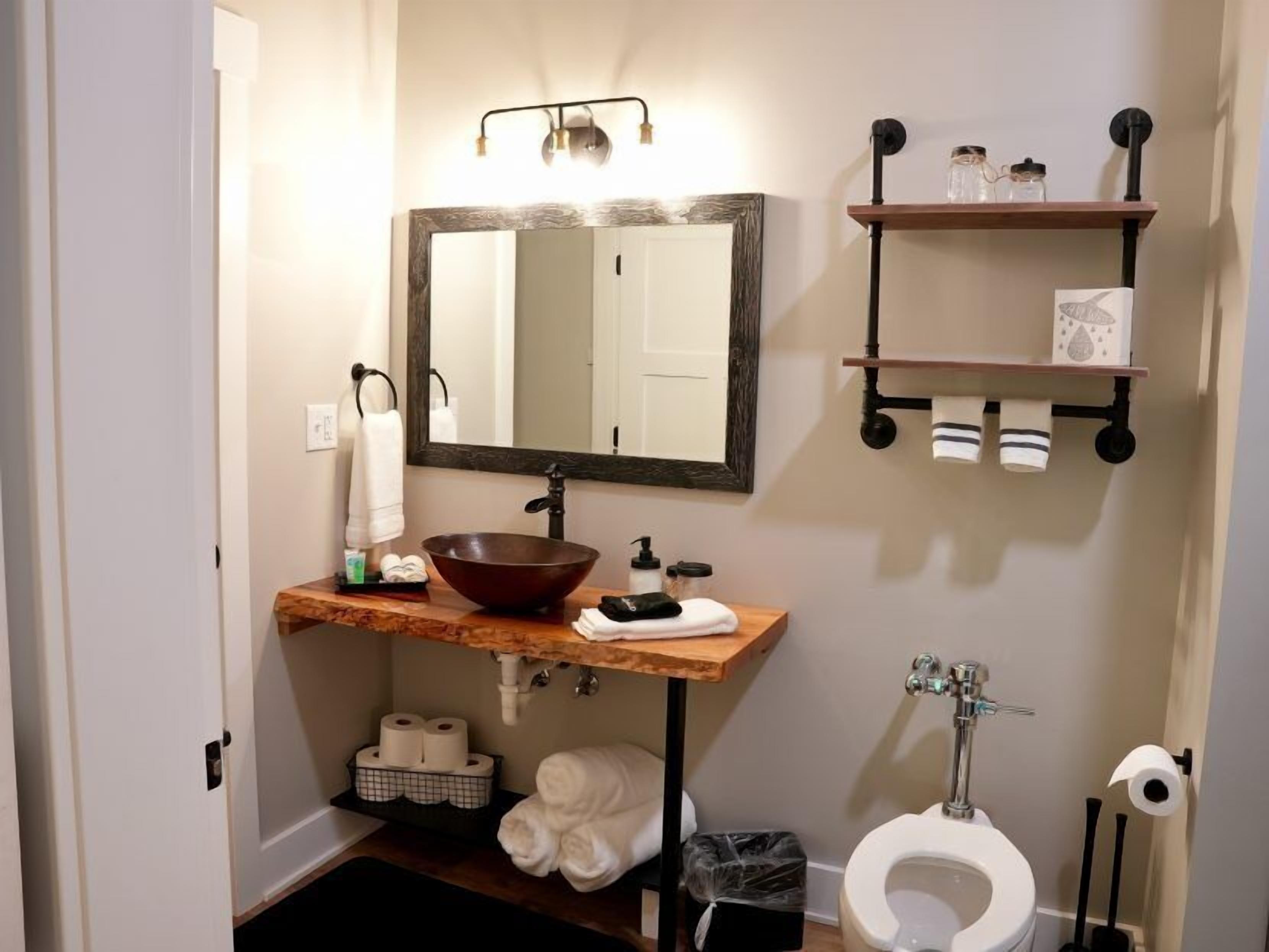 A bathroom with a wooden vanity, a large mirror, a white toilet, and a shelf with toiletries.