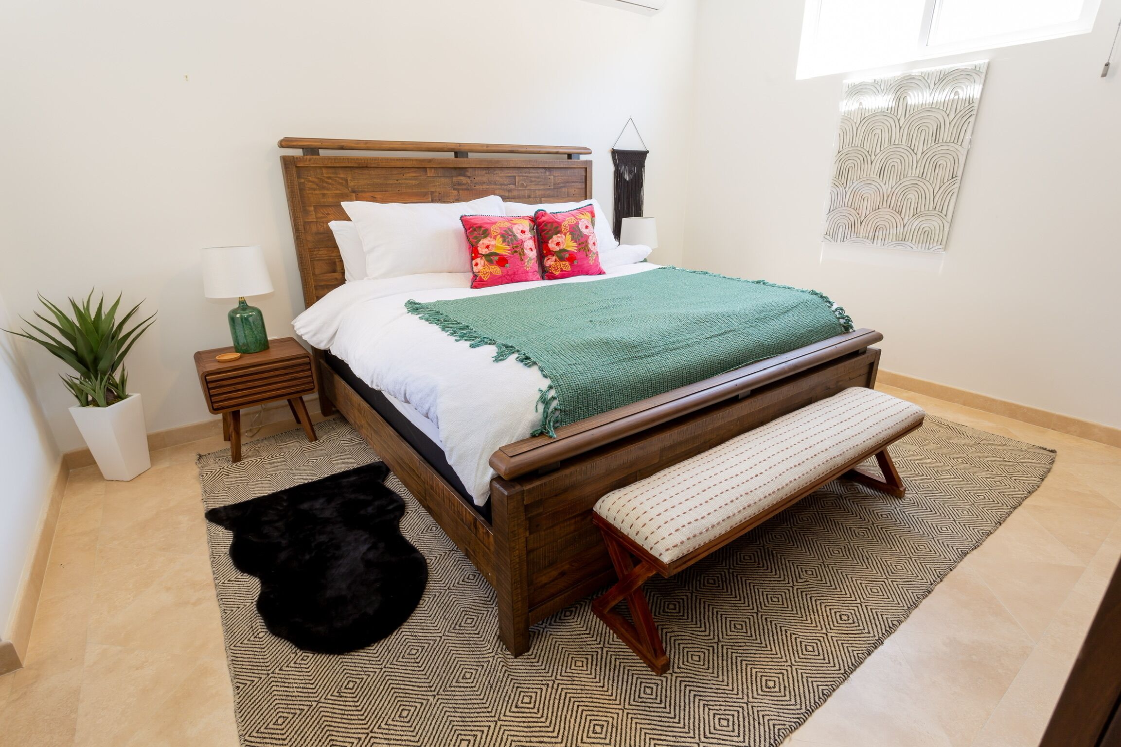 A neatly arranged bedroom with a wooden bed, a green throw blanket, a bench, a potted plant, and a patterned rug.